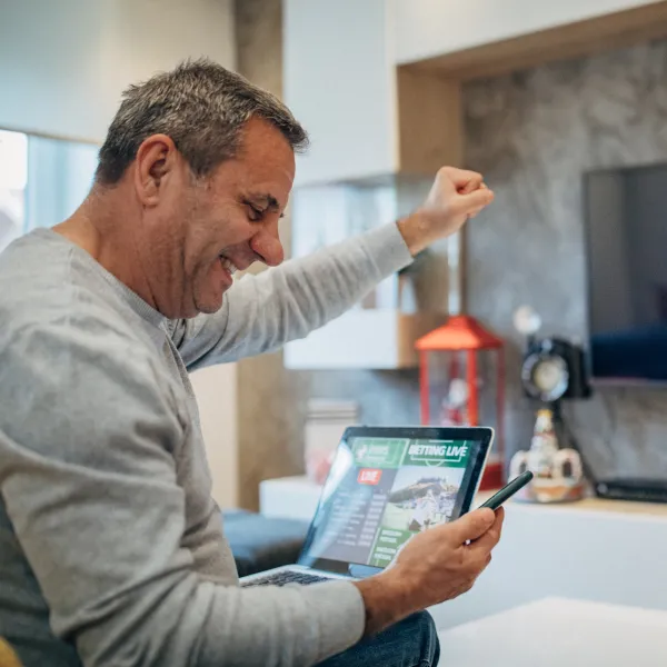 A man sitting on a couch celebrates with a raised fist while looking at his phone and laptop, both displaying a sports betting website.