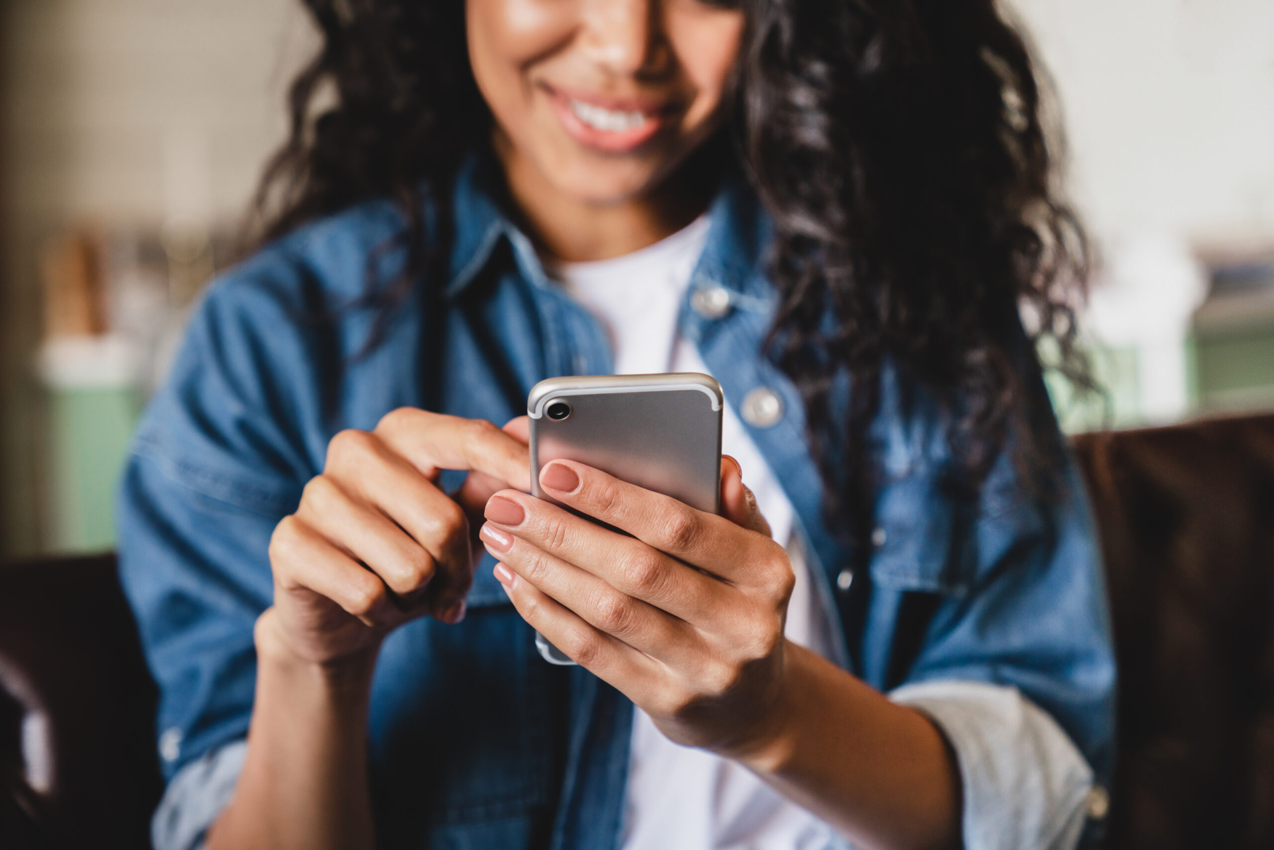 A woman with curly hair, wearing a denim jacket, smiles while using a smartphone, holding it with one hand and tapping the screen with the other. The background is softly blurred.