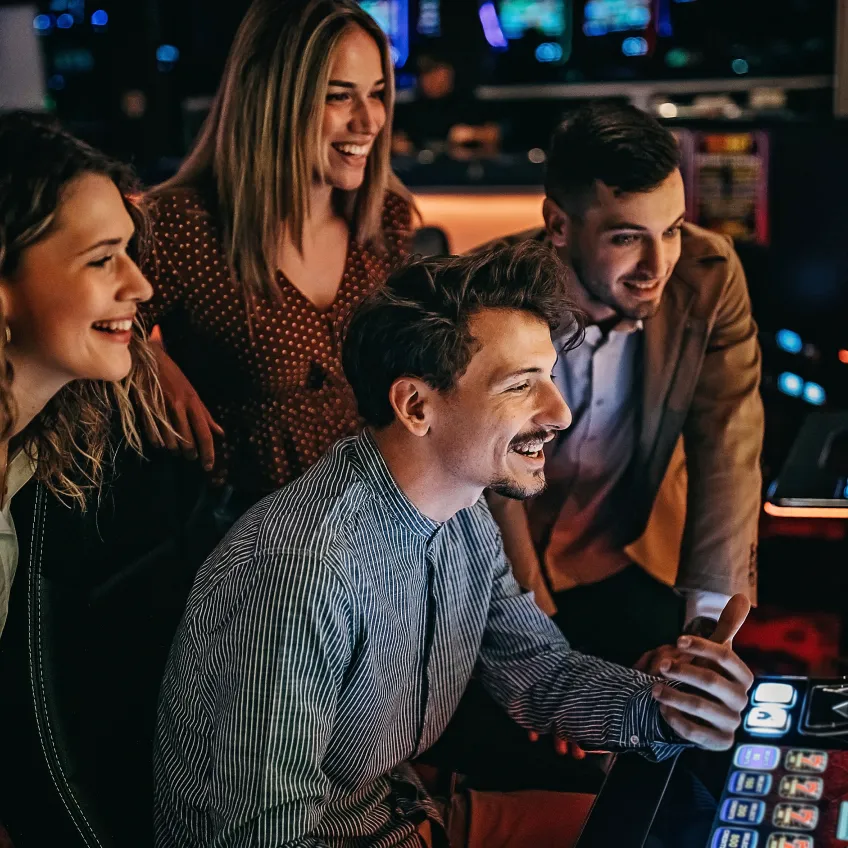 Four young adults, two women and two men, smile and watch excitedly as one man plays a slot machine in a lively, colorful casino setting.