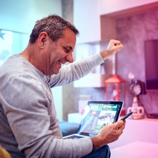 A man sitting on a couch smiles and raises his fist in celebration while looking at his smartphone, with a laptop showing a sports betting website on his lap.