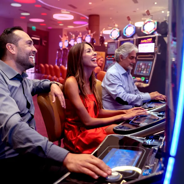 Three people sit side by side at slot machines in a casino, smiling and focused on playing. The atmosphere is lively, with neon lights and more machines in the background.