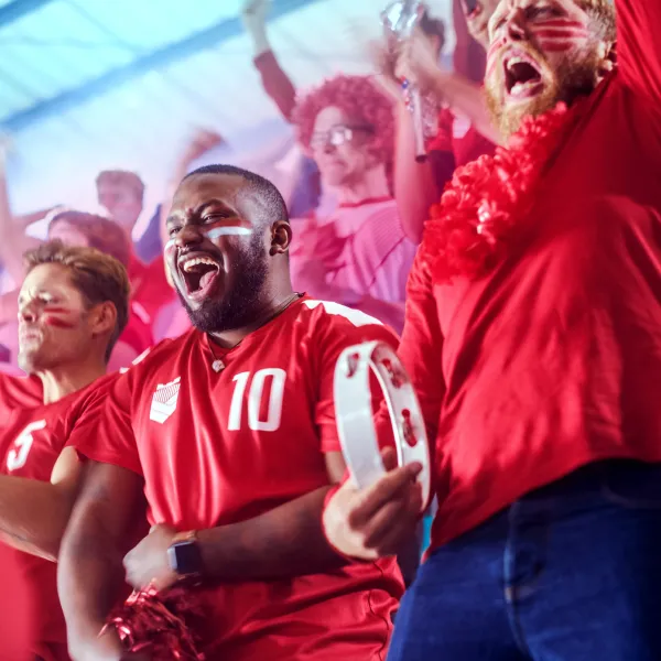 A group of enthusiastic sports fans in red shirts with face paint cheer and celebrate at a stadium. One person holds a tambourine, and everyone appears energetic and joyful.