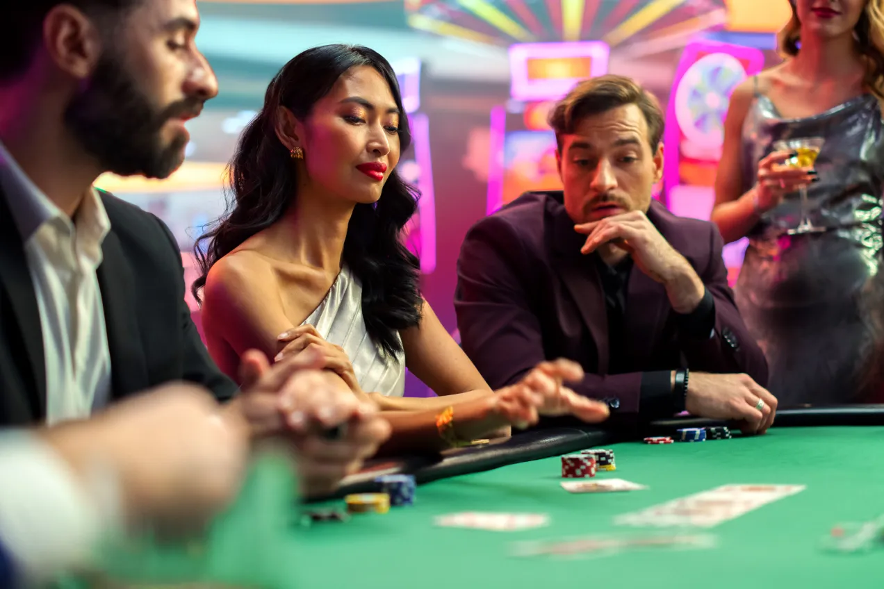 Three people in formal attire sit at a casino poker table, focused on the game. Poker chips and cards are on the green felt table, with colorful lights and slot machines in the background.